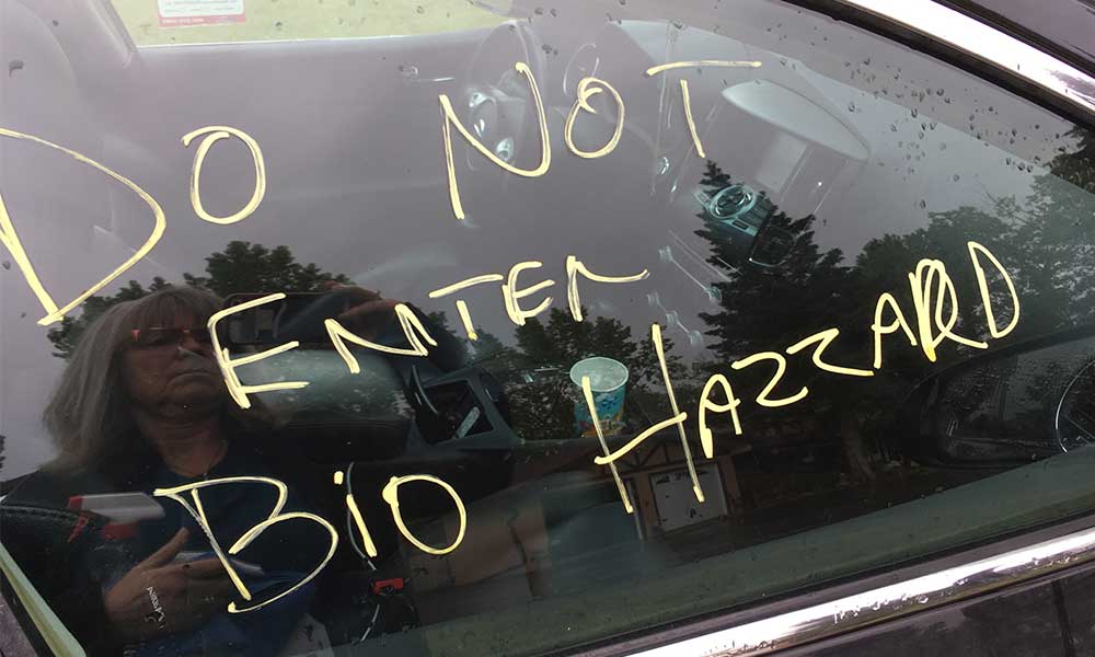 Car window with a handwritten message in yellow that reads 'Do Not Enter Bio Hazard,' with a reflection of a person in sunglasses and trees in the background.