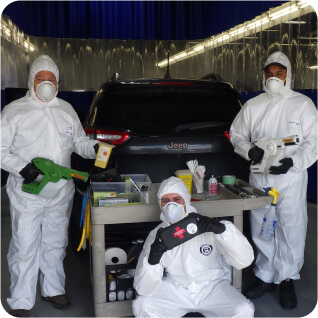 Three hazmat team members in protective suits and masks pose with cleaning equipment and supplies in front of a Jeep, ready for vehicle sanitization.
